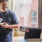 Lean Startup - Happy adult bearded man in home outfit leaning on table with modern laptop and drinking coffee during break in freelance work on sunny day