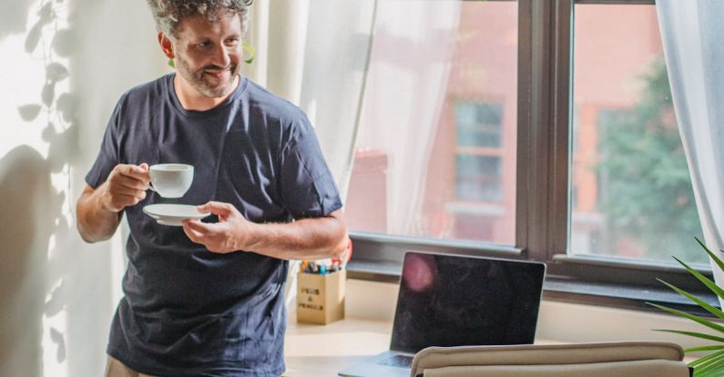 Lean Startup - Happy adult bearded man in home outfit leaning on table with modern laptop and drinking coffee during break in freelance work on sunny day