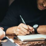 Cash Flow Analysis - Crop elegant man taking notes in journal while working at desk with coins and piggybank in lamplight