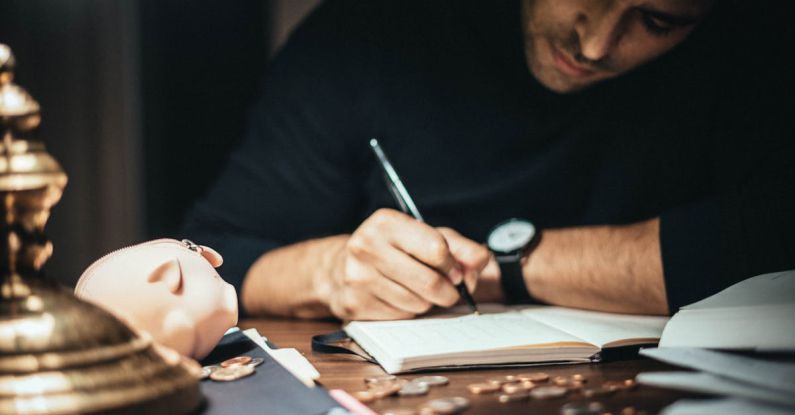 Cash Flow Analysis - Crop elegant man taking notes in journal while working at desk with coins and piggybank in lamplight